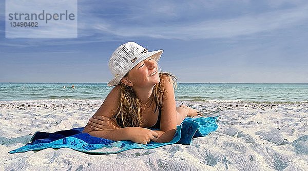 Mädchen mit Sonnenhut auf einem blauen Strandtuch liegend am Meer an einem weißen Sandstrand