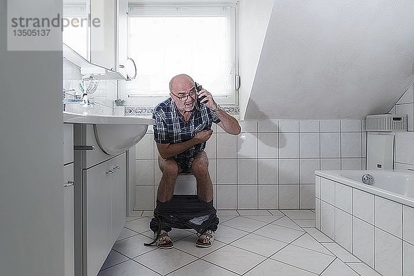 Älterer Mann sitzt auf der Toilette und spricht in ein Smartphone  Deutschland  Europa