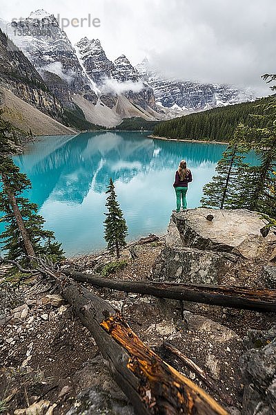 Junge Frau steht vor einem See und blickt in eine Berglandschaft  Wolken hängen zwischen Berggipfeln  Spiegelung im türkisfarbenen See  Moraine Lake  Valley of the Ten Peaks  Rocky Mountains  Banff National Park  Provinz Alberta  Kanada  Nordamerika