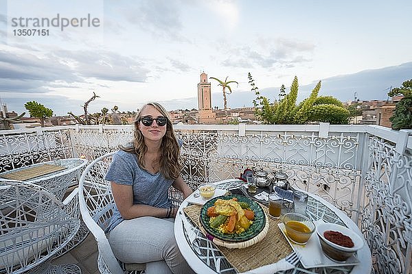 Junge Frau auf einer Dachterrasse im Restaurant  marokkanisches Essen  Tagine  Blick auf die Altstadt  Moschee mit Minarett  Abendstimmung  Marrakech  Marokko  Afrika