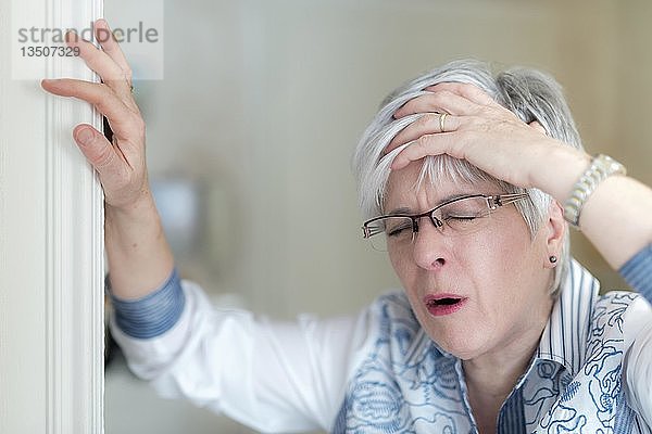 Ältere Frau mit Kopfschmerzen hält sich die Hand auf die Stirn und stützt sich auf den Türrahmen  Deutschland  Europa