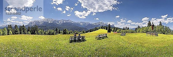 Wanderer entspannen sich auf einer Bank inmitten einer Blumenwiese mit Panoramablick auf das Wettersteinmassiv mit den Alpen und der Zugspitze  Wamberg  Oberbayern  Bayern  Deutschland  Europa