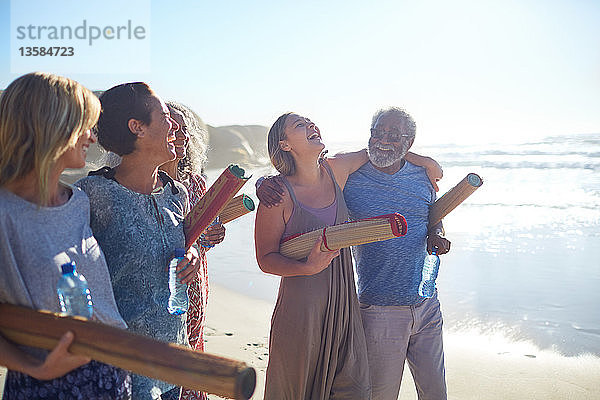 Glückliche Freunde mit Yogamatten  die sich am sonnigen Strand während eines Yoga-Retreats treffen