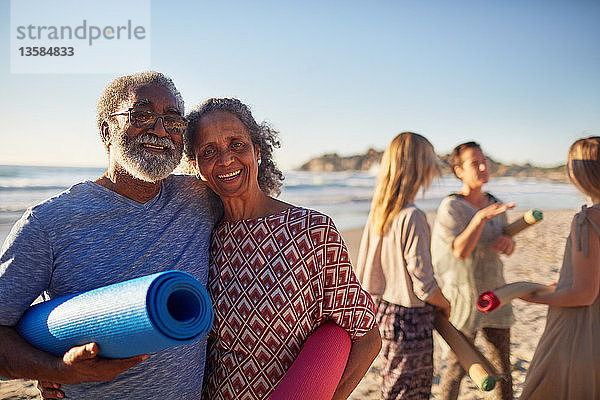 Portrait glückliches älteres Paar mit Yogamatten am sonnigen Strand während eines Yoga-Retreats