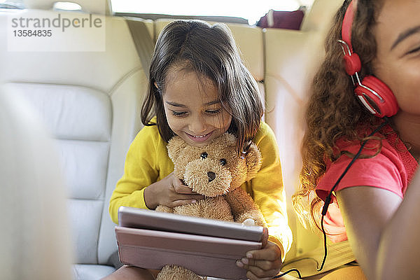 Niedliches Mädchen mit Teddybär  das ein digitales Tablet auf dem Rücksitz eines Autos benutzt