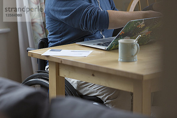 Junge Frau im Rollstuhl mit Laptop am Esstisch