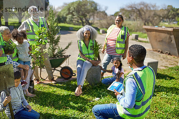 Mann mit digitalem Tablet führt Freiwillige an  die in einem sonnigen Park Bäume pflanzen