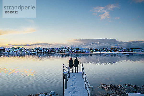 Händchen haltendes Paar am Rande eines verschneiten Docks mit Blick auf ein Dorf am Wasser  Reine  Lofoten  Norwegen