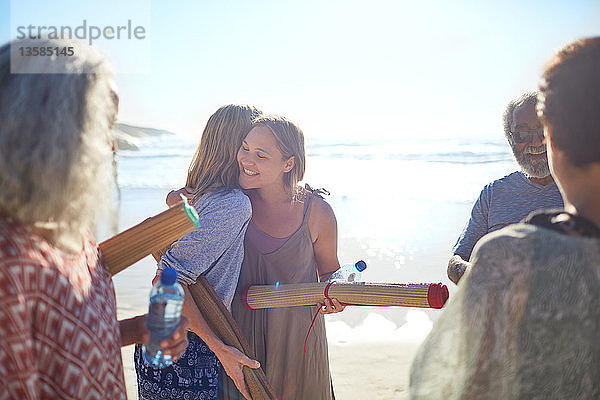 Freundinnen mit Yogamatten umarmen sich am sonnigen Strand während eines Yoga-Retreats