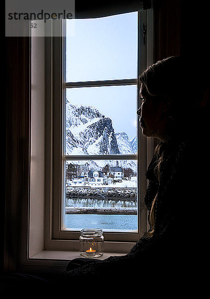 Nachdenkliche Frau schaut aus dem Fenster auf einen verschneiten Berg  Reine  Lofoten  Norwegen