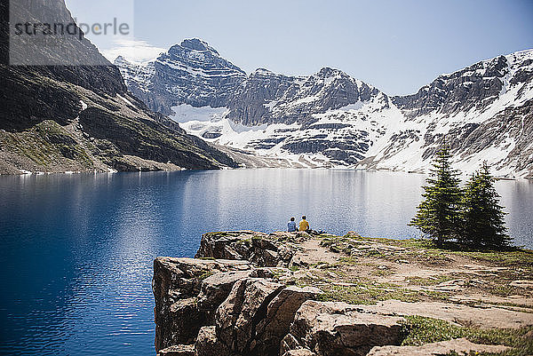 Paar sitzt auf einer Klippe mit Blick auf ruhige  sonnige Berge und einen See  Yoho Park  British Columbia  Kanada