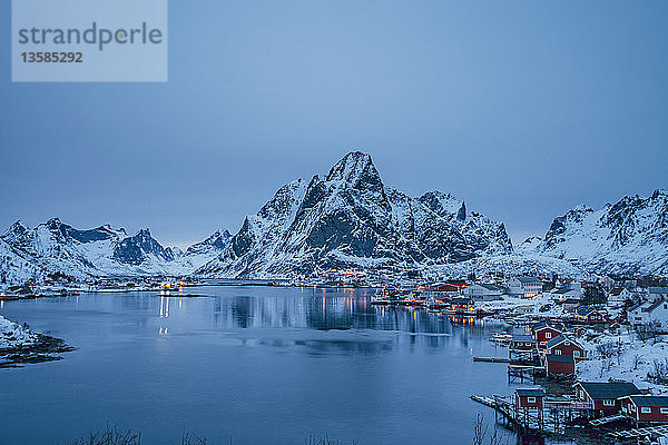 Ruhiger Blick auf das schneebedeckte Fischerdorf am Wasser und die Berge bei Nacht  Reine  Lofoten Inseln  Norwegen