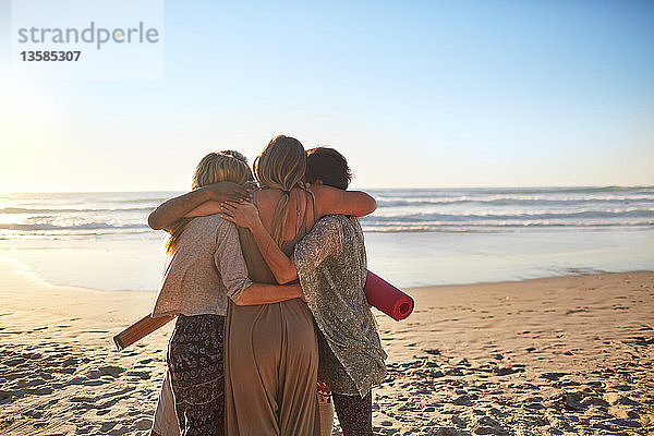Freundinnen mit Yogamatten umarmen sich am sonnigen Strand