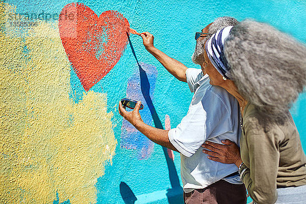 Älteres Paar malt herzförmiges Wandgemälde an eine sonnige Wand