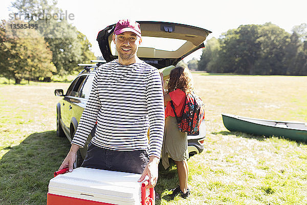 Porträt eines lächelnden Mannes  der eine Camping-Kühlbox trägt und sein Auto in einem sonnigen Feld entlädt