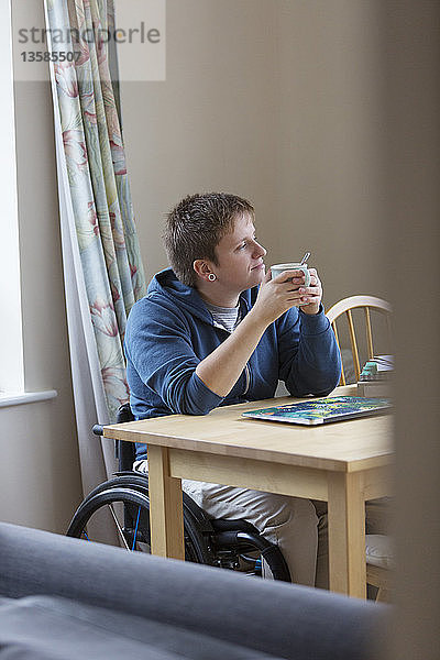 Nachdenkliche junge Frau im Rollstuhl trinkt Tee am Esstisch