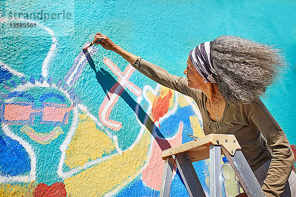Eine ältere Frau malt ein lebendiges Wandgemälde an eine sonnige Wand