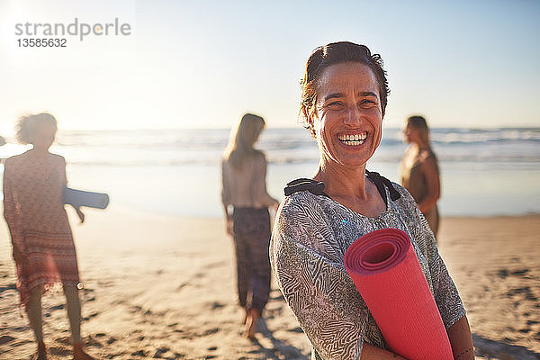 Portrait glückliche Frau mit Yogamatte am sonnigen Strand während eines Yoga-Retreats