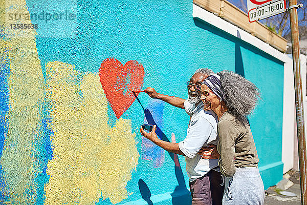 Glückliches älteres Paar malt herzförmige Wandbilder an eine sonnige Wand