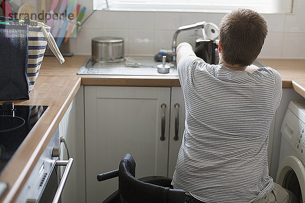 Junge Frau im Rollstuhl füllt einen Wasserkocher für Tee an der Küchenspüle einer Wohnung