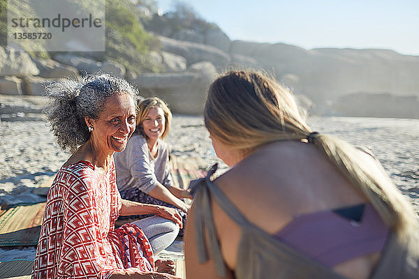 Lächelnde Freundinnen unterhalten sich am sonnigen Strand während eines Yoga-Retreats