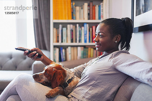 Junge Frau und Hund entspannen sich auf dem Wohnzimmersofa und sehen fern