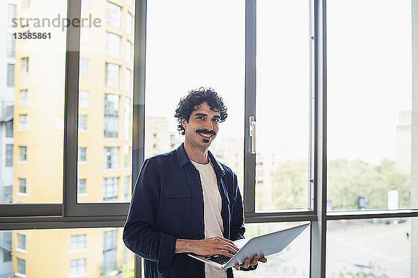Porträt eines selbstbewussten Mannes  der einen Laptop am Fenster einer städtischen Wohnung benutzt