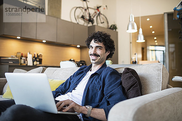 Porträt eines lächelnden Mannes mit Laptop auf dem Sofa einer Wohnung