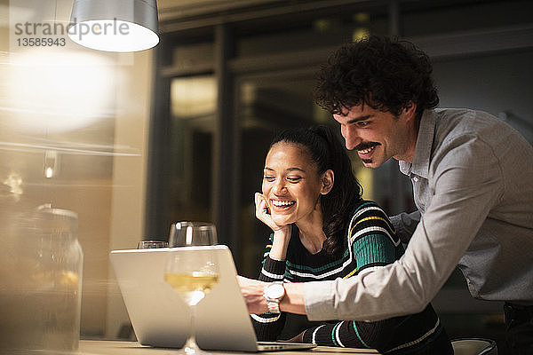 Glückliches Paar  das abends zu Hause einen Laptop benutzt und Weißwein trinkt