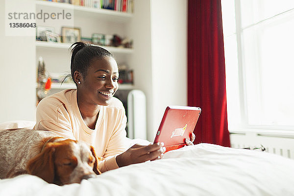 Lächelnde junge Frau mit digitalem Tablet neben einem schlafenden Hund auf dem Bett