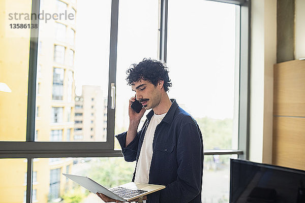 Mann benutzt Laptop und spricht mit Smartphone am Fenster einer städtischen Wohnung