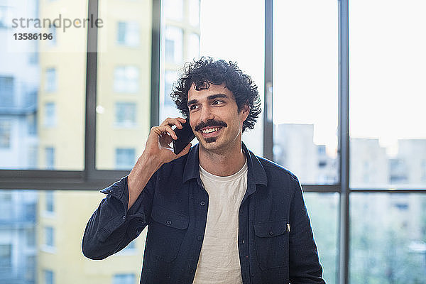 Lächelnder Mann  der am Fenster einer Stadtwohnung mit seinem Smartphone telefoniert