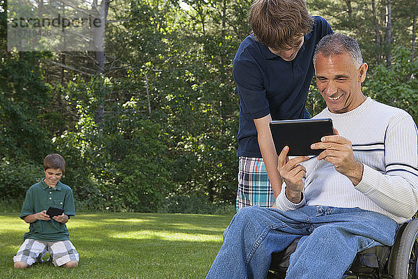 Mann mit Rückenmarksverletzung im Rollstuhl mit seinen Söhnen beim Lesen eines Tablets