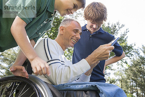 Mann mit Rückenmarksverletzung im Rollstuhl mit seinen Söhnen beim Lesen eines Tablets