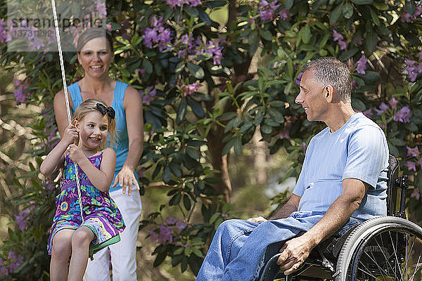 Mann mit Rückenmarksverletzung im Rollstuhl beobachtet seine Tochter auf der Schaukel zusammen mit seiner Frau