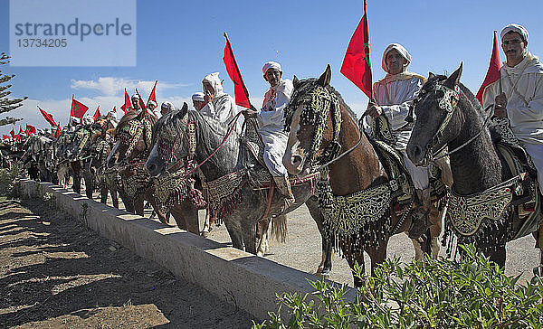 Männer auf Pferden in traditionellen weißen Gewändern schwenken die rote Nationalflagge  Essaouira  Marokko