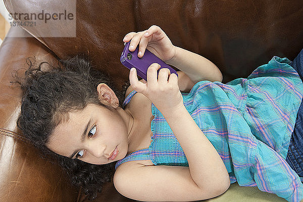 Hispanisches Mädchen  das auf einer Couch liegt und sich eine Textnachricht auf einem Mobiltelefon ansieht