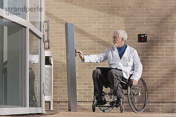 Arzt mit Muskeldystrophie im Rollstuhl am Krankenhauseingang drückt auf den Knopf für eine zugängliche Tür