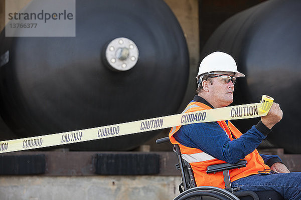 Gebäudetechniker im Rollstuhl beim Ziehen eines Absperrbandes vor einem Chemielagertank