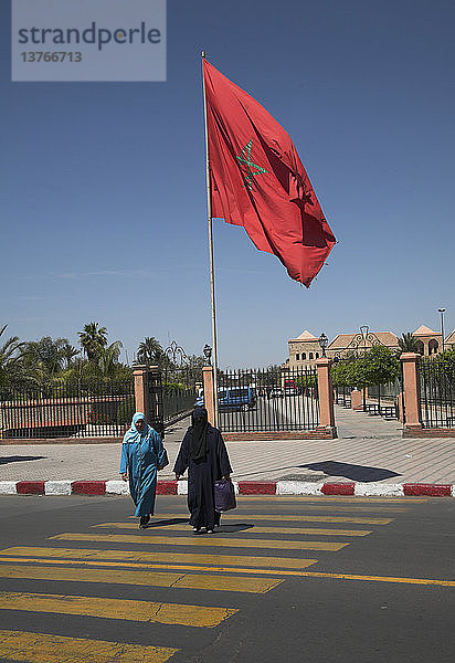 Große rote marokkanische Flagge weht  während zwei muslimische Frauen die Straße überqueren Marrakesch  Marokko  Nordafrika