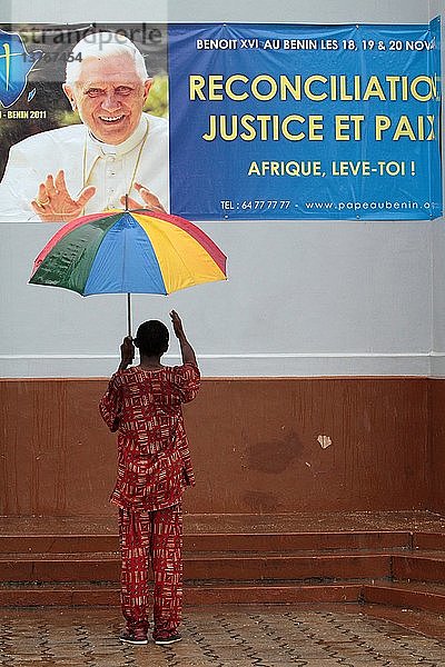 Afrikaner  hinten  trägt einen Regenschirm vor einem Plakat  das den Besuch von Papst Benedikt XVI. in Benin ankündigt.