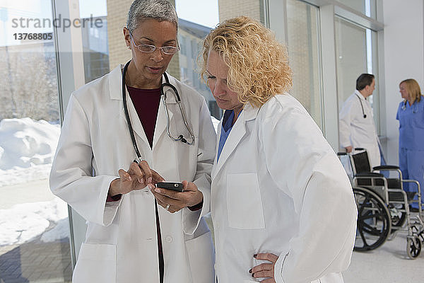 Zwei Ärztinnen lesen eine Textnachricht auf einem Mobiltelefon  während ihre Kollegen im Hintergrund stehen