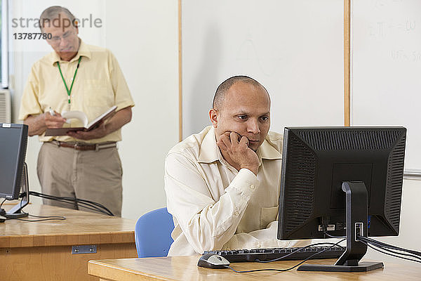 Ingenieurstudent studiert Computerbildschirm in einem Klassenzimmer mit Professor im Hintergrund
