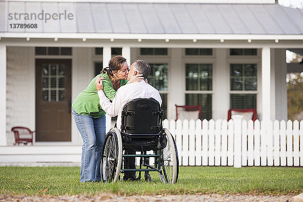 Pärchen küsst sich vor ihrem neuen Haus  während er im Rollstuhl sitzt