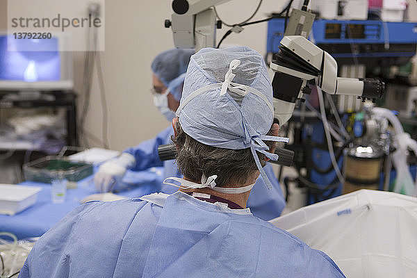 Arzt schaut während eines chirurgischen Eingriffs in ein Mikroskop  während ein Operationstechniker das nächste Instrument nimmt