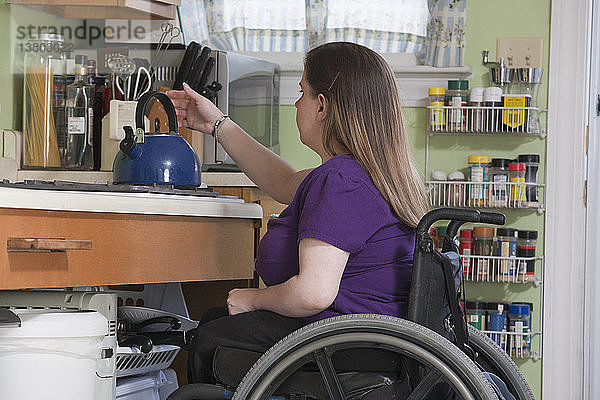 Frau mit Spina Bifida im Rollstuhl beim Aufstellen eines Wasserkochers in der barrierefreien Küche