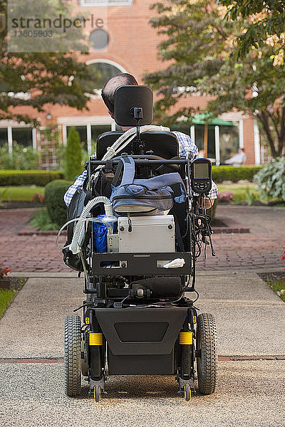 Rückansicht eines Mannes mit Duchenne-Muskeldystrophie in einem motorisierten Rollstuhl auf der Straße