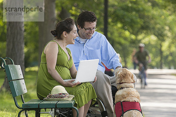 Schwangere Frau mit Laptop sitzt im Park mit Ehemann im Rollstuhl und Diensthund