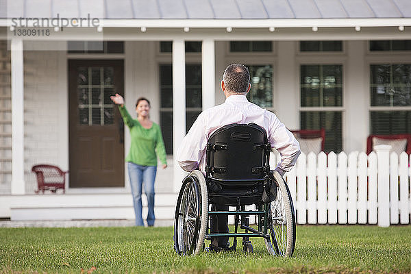 Frau winkt von ihrem Haus aus ihrem Mann im Rollstuhl zu  der eine Rückenmarksverletzung hat