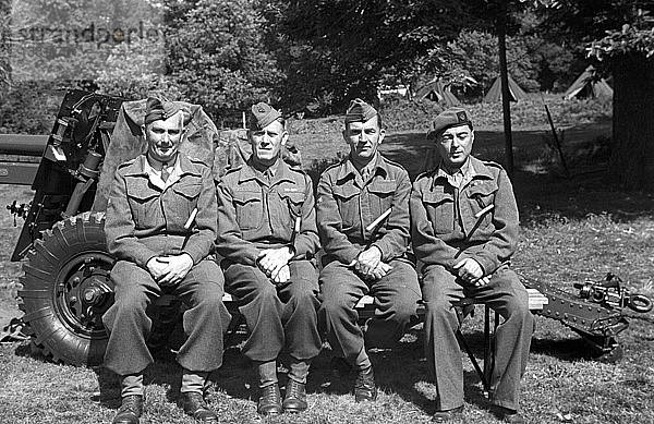 Vier Armeeoffiziere  die auf einem Artilleriegeschütz sitzen und mit Stöcken posieren. In der Ferne sind Zelte zu sehen. Männer in Uniform  Soldaten.